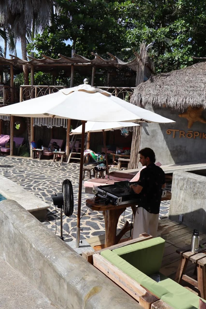 Daytime set at an open-air beach club under thatched roofs and palms